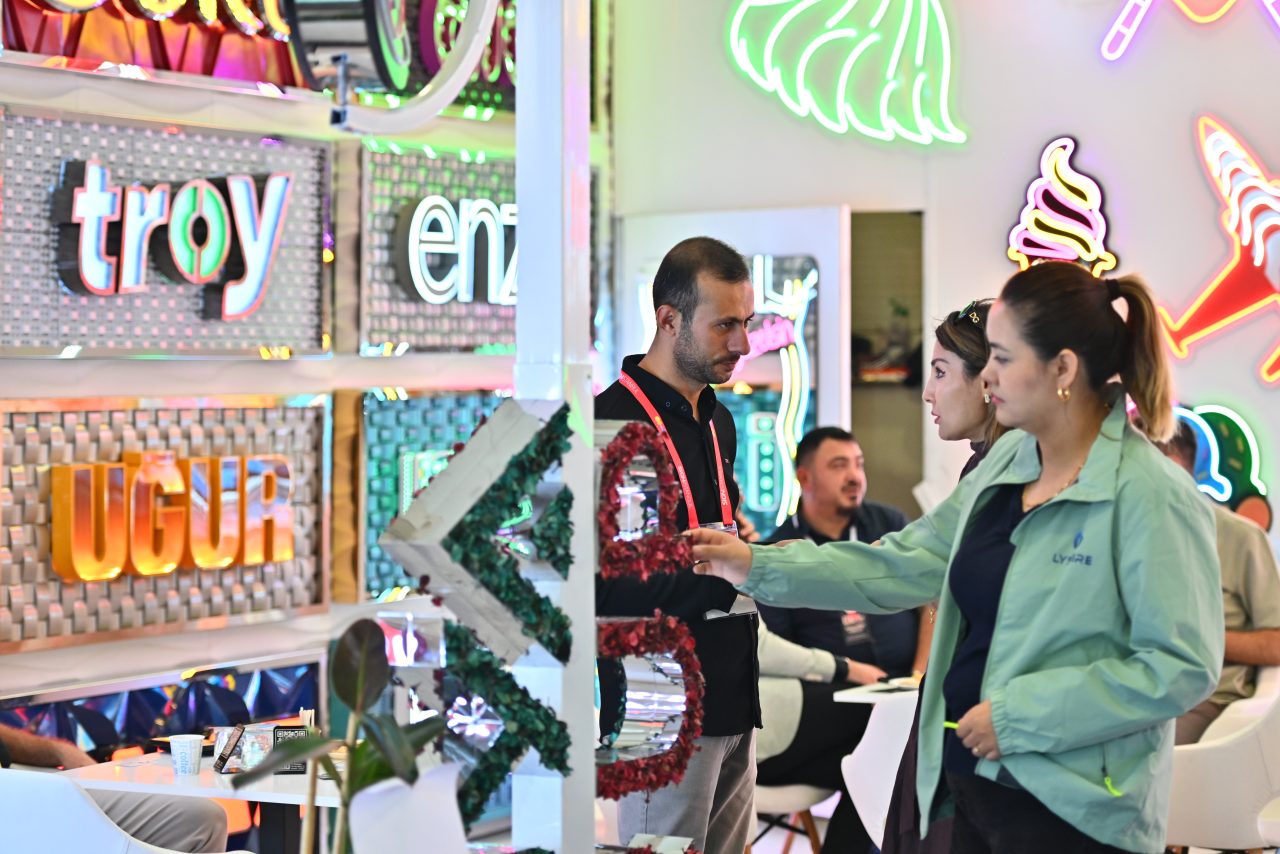 Bir grup ziyaretçi, sergi standında oturmuş, birinin konuşmasını dikkatle dinliyor.